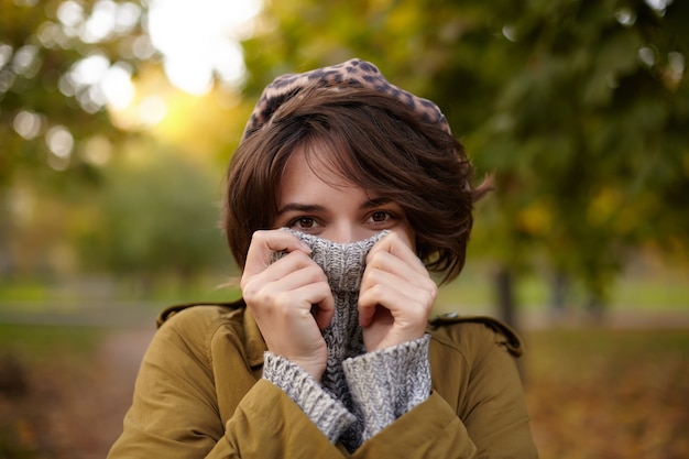 Free photo outdoor photo of attractive brown-eyed young brunette female with natural makeup wearing stylish clothes while posing over blurred park and hiding her face